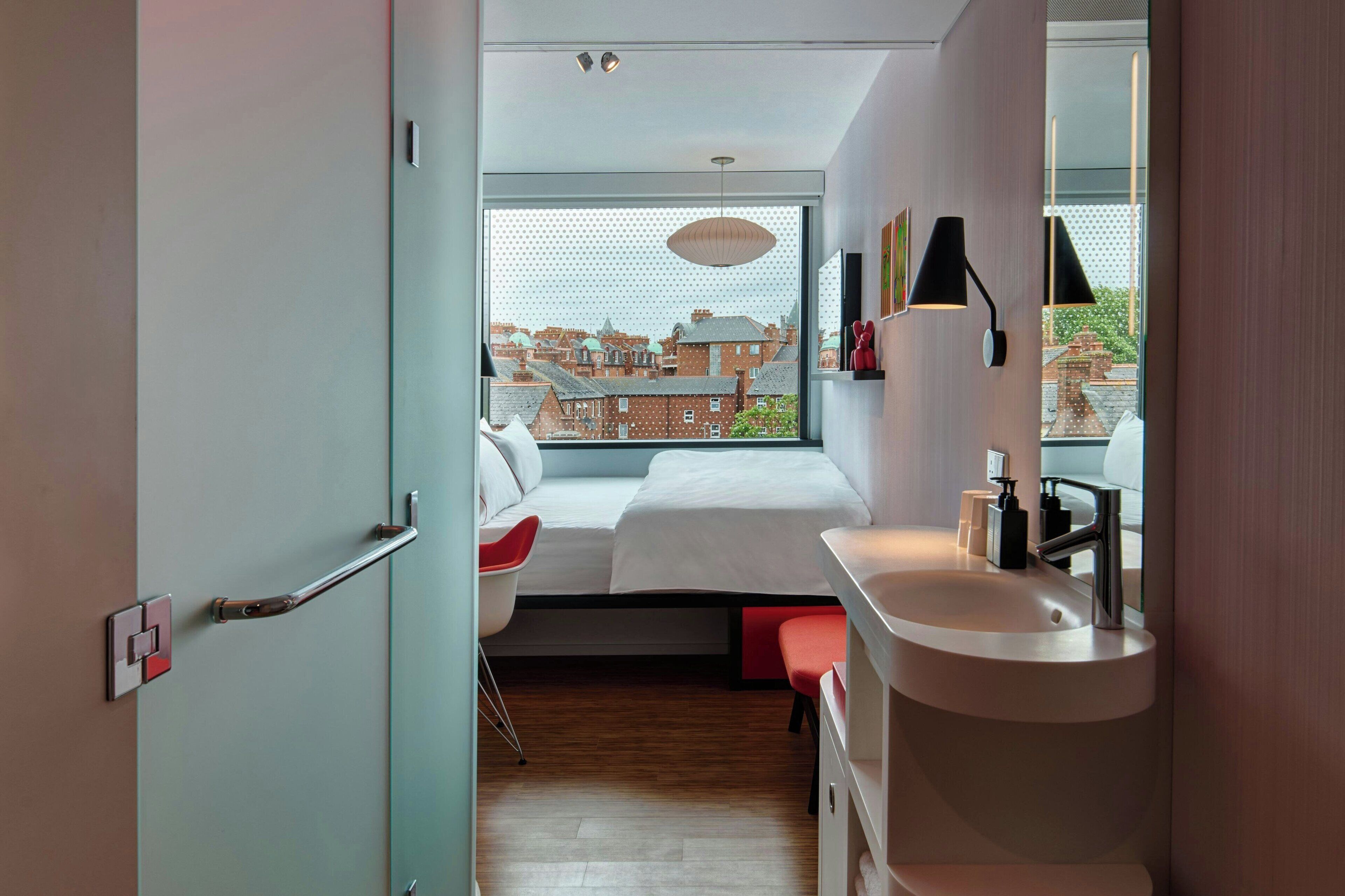 Een moderne slaapkamer met een groot raam, een bed, een nachtkastje, een stoel, en een badkamer met een wastafel en spiegel.