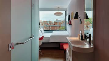 Une chambre moderne avec une grande fenêtre, un lit, une table de chevet, une chaise et une salle de bain équipée d’un lavabo et d’un miroir.