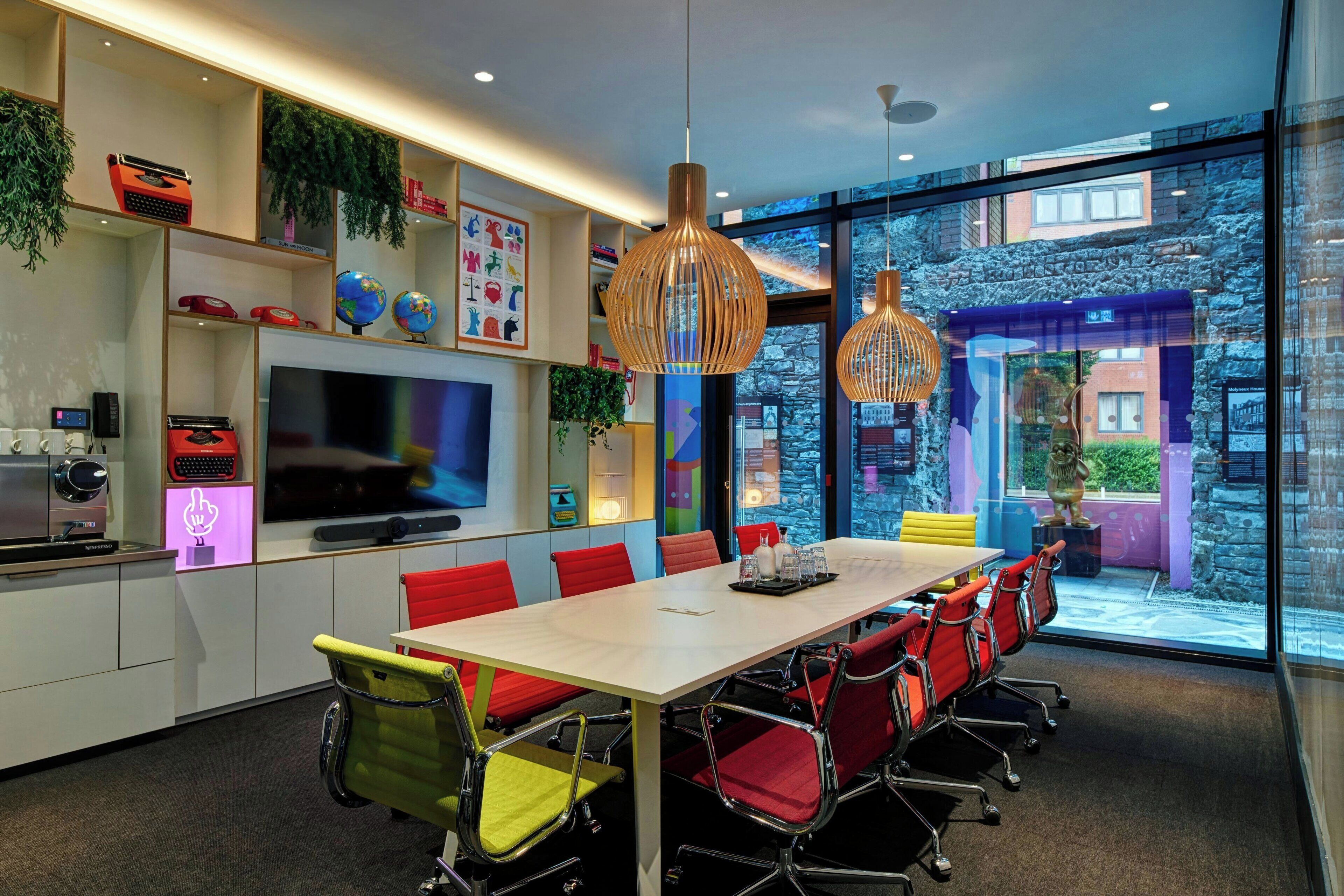 A modern meeting room with a large table, red chairs, a TV, and shelves with decorative items.