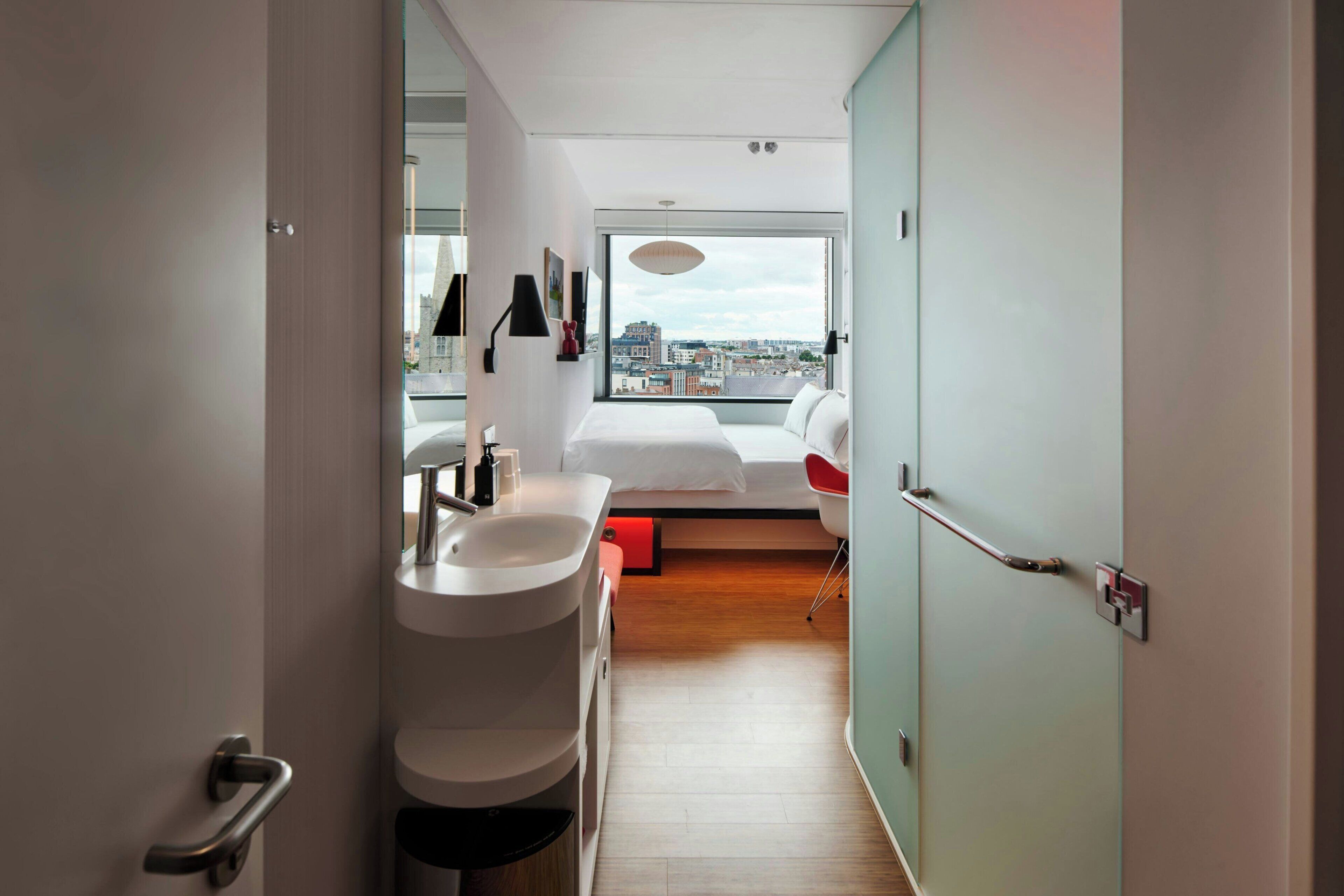 Una camera d'albergo moderna con un'ampia finestra, un letto, un bagno con un lavandino e una poltrona rossa.
