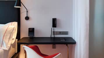 A modern hotel room with a neatly made bed, a desk with a lamp, a chair, and a power outlet.