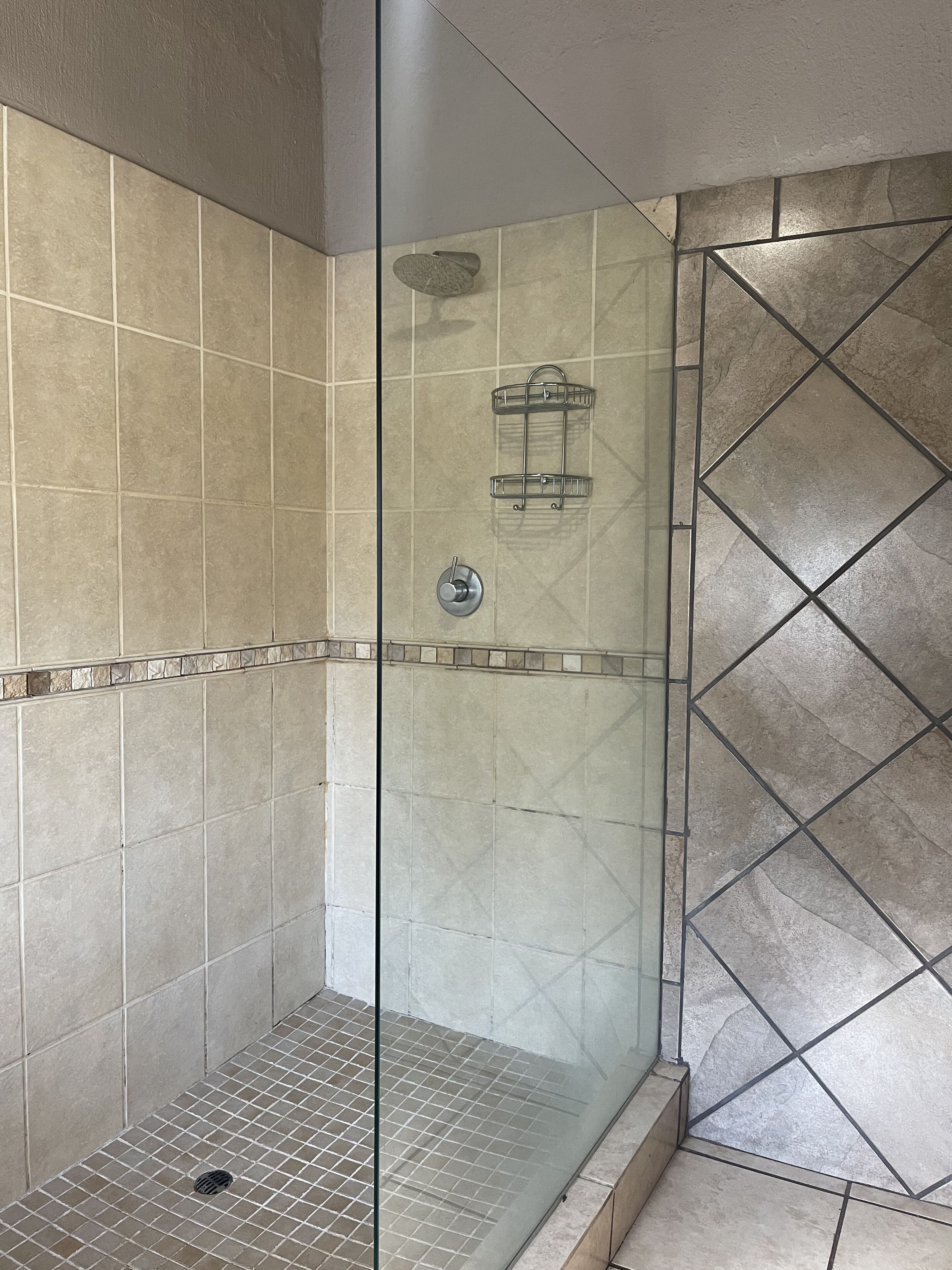 Een douche met glazen wand, betegelde vloer en wanden, een douchekop en een metalen plank.