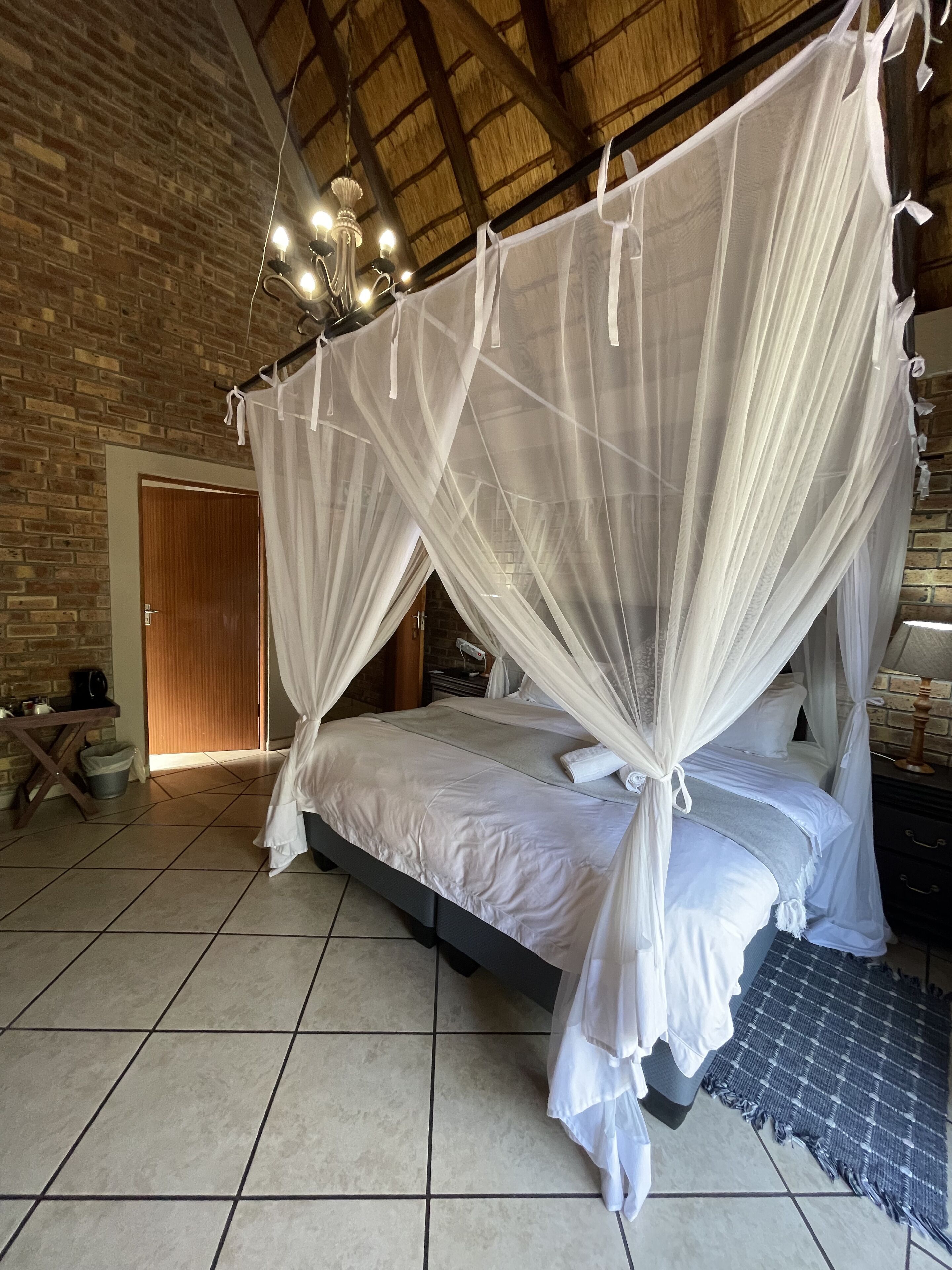 Quarto com cama sob um dossel branco, porta de madeira, parede de tijolos e chão de azulejos.