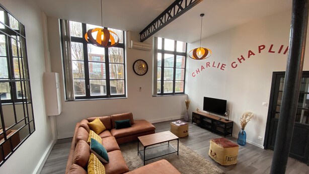 Un salon moderne avec un canapé d’angle, une table basse et un téléviseur. La pièce est dotée de grandes fenêtres, d’une cheminée et d’un escalier avec des poutres apparentes.