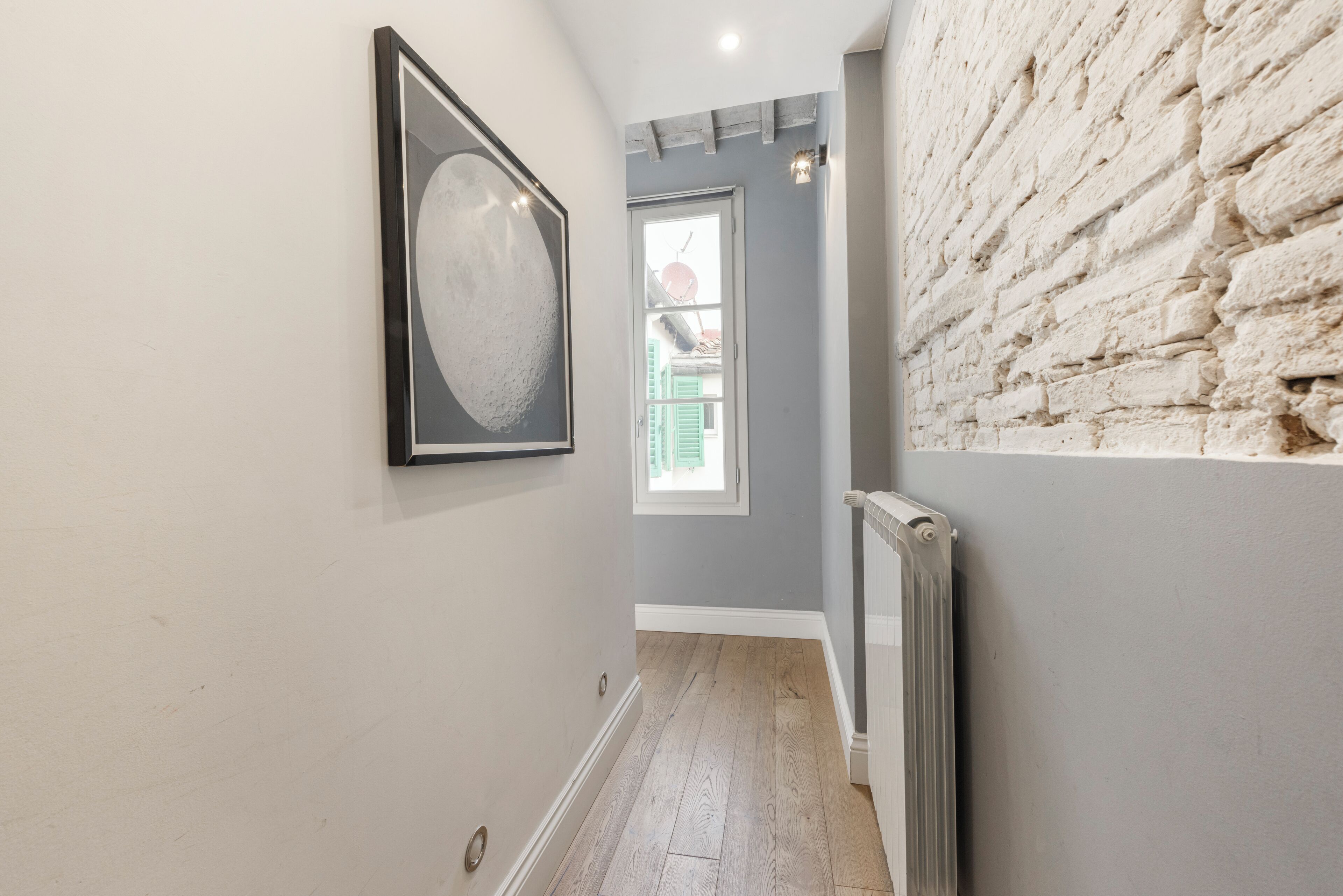 A hallway with a wooden floor, a framed picture on the wall, a window with shutters, and a brick wall.