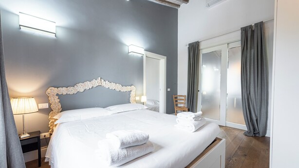 A neatly made bed with white linens and towels, a bedside table with a lamp, and a chair near a sliding glass door. - Central Nest by MMega (Florence)