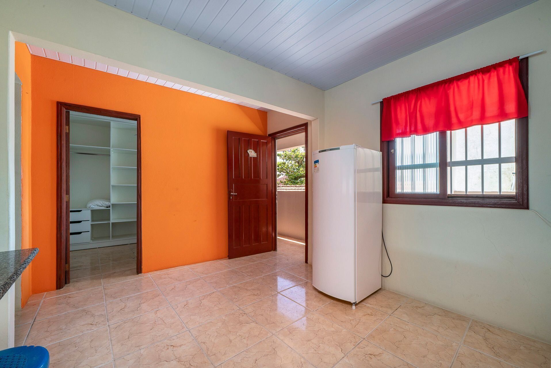 A room with a tiled floor, a white refrigerator, a red curtain over a window, and a wooden door leading to a closet.
