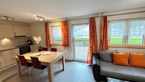 Eine moderne Küche mit weißen Schränken und einem Essbereich mit Holztisch und -stühlen. Ein Sofa mit orangefarbenen Kissen und Blick auf einen Garten durch die Fenster.
