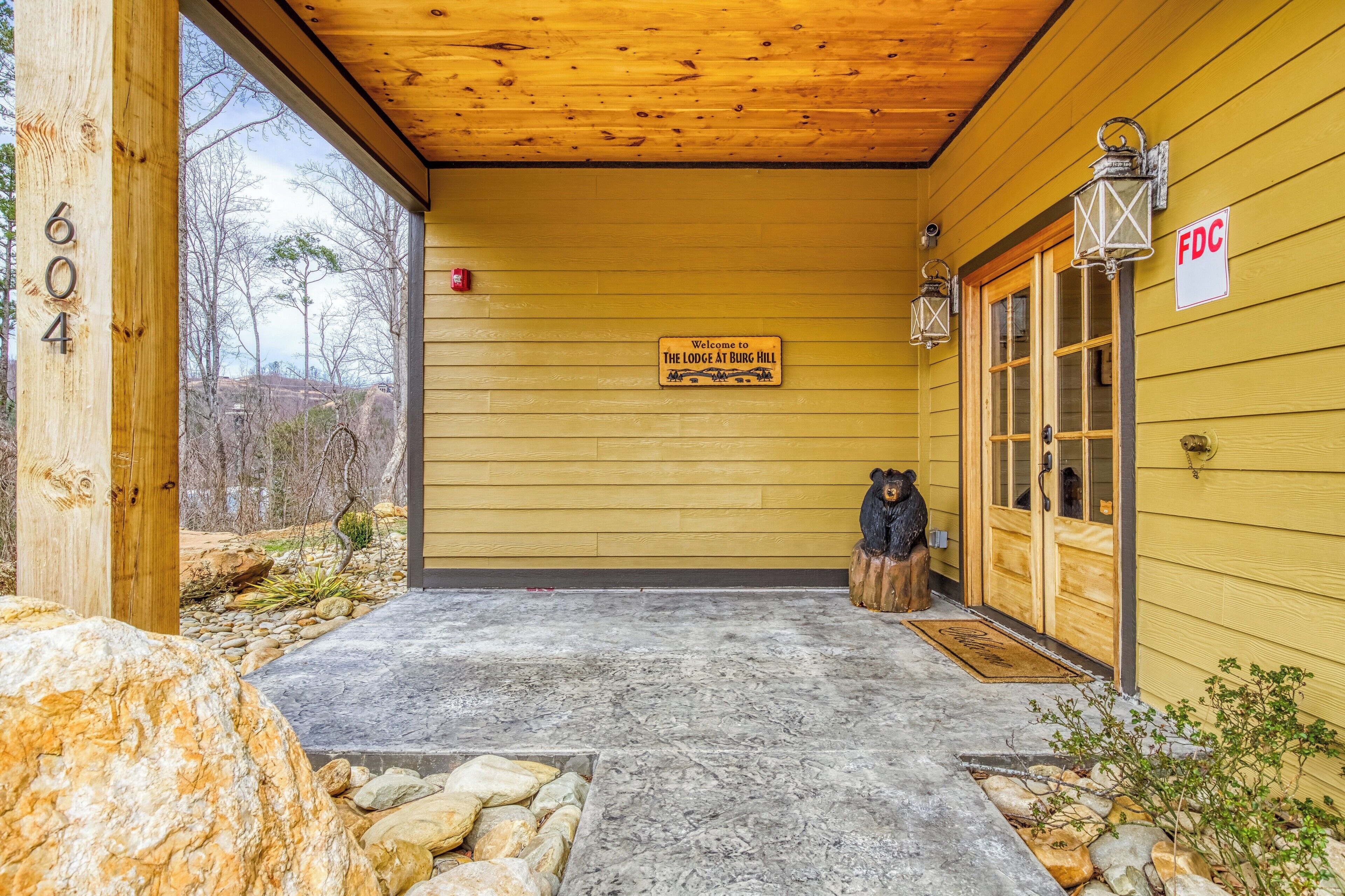A wooden lodge entrance with a welcome sign, a bear statue, and a stone pathway leading to the door.
