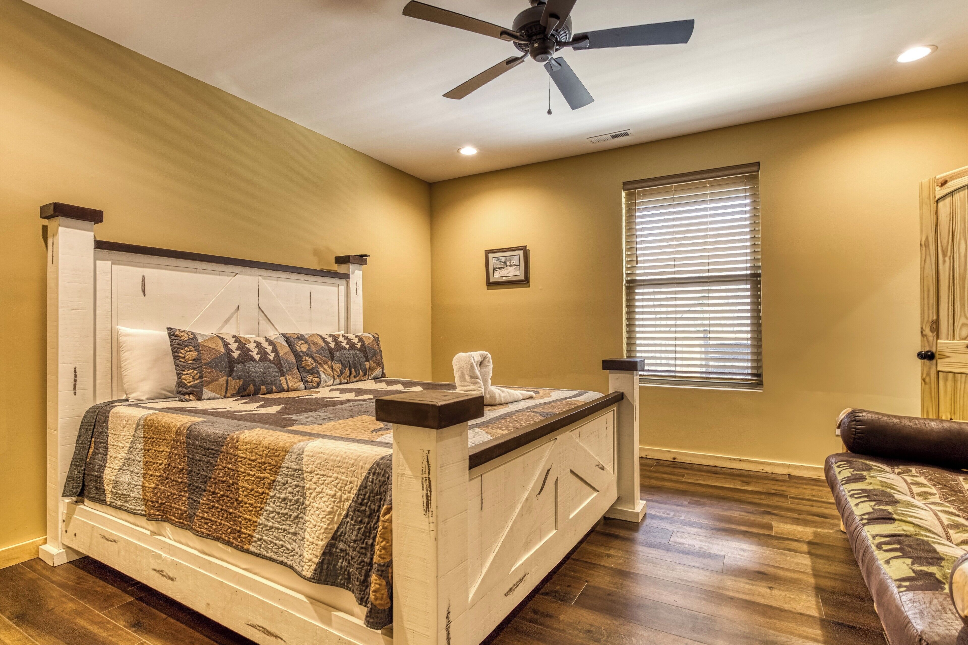 Une chambre à coucher avec un lit, un ventilateur de plafond, une fenêtre avec des stores et une porte en bois.