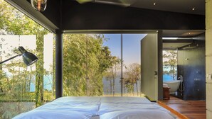 A modern bedroom with a large bed, a desk lamp, and a view of a garden and water body. - Casa Ceibo - Private Villa with Pool (Puntarenas Province)