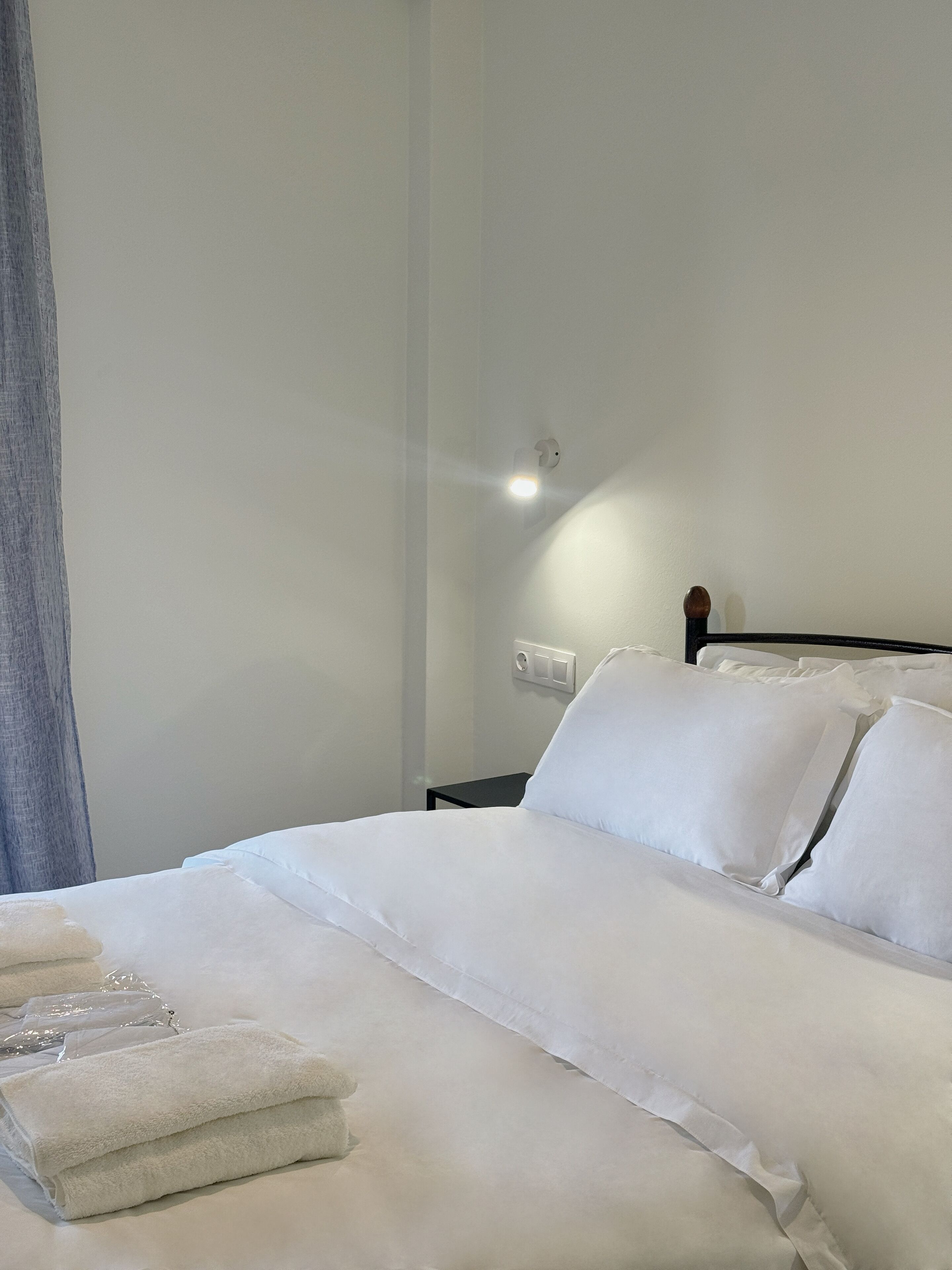 Una cama bien tendida con sábanas y almohadas blancas, una mesita de noche con lámpara, y un aplique en la pared.