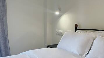 Una cama bien tendida con sábanas y almohadas blancas, una mesita de noche con lámpara, y un aplique en la pared.