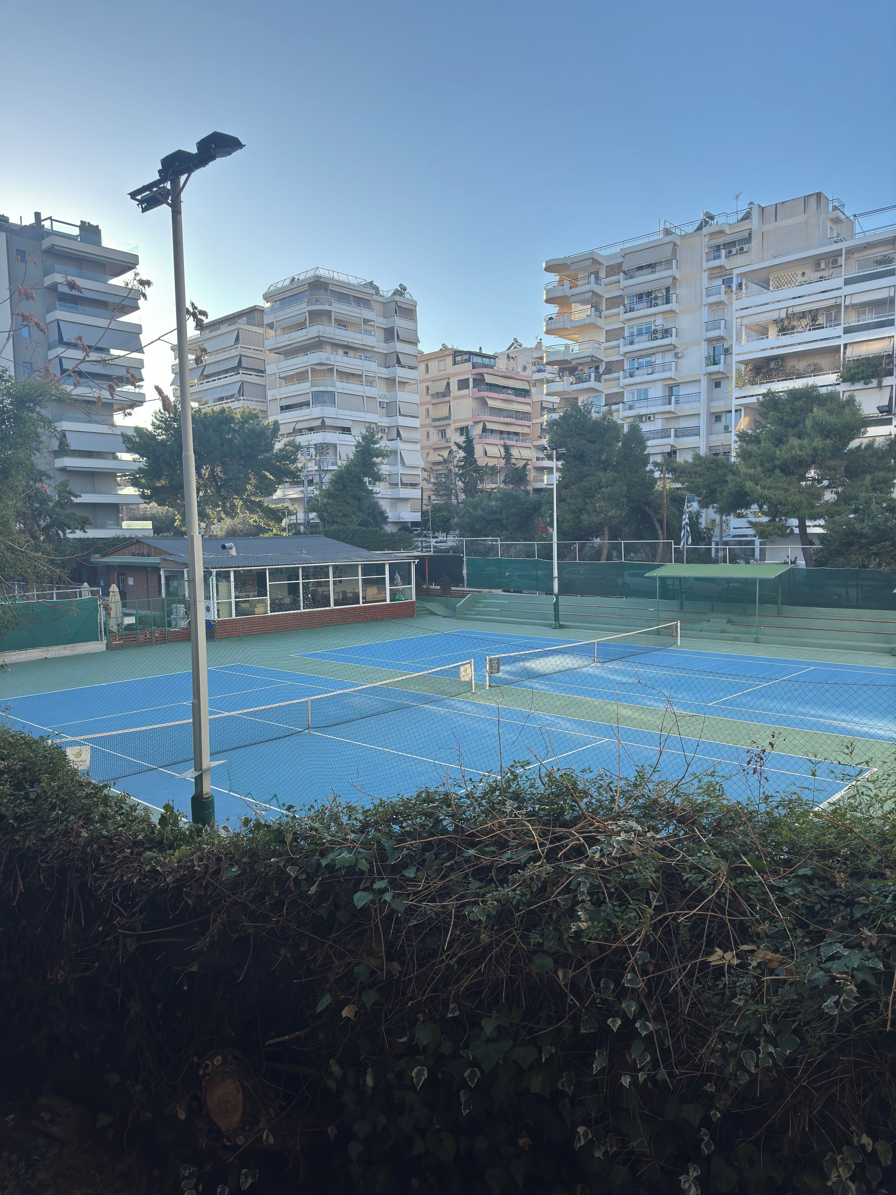 A tennis court with a net, surrounded by a fence, and buildings in the background.