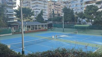 A tennis court with a net, surrounded by a fence, and buildings in the background.