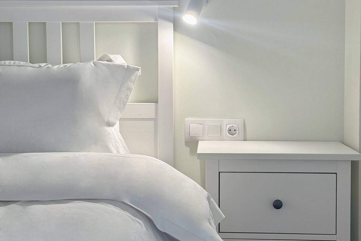 A neatly made bed with white linens and pillows, a white nightstand with a drawer, and a wall-mounted light fixture.
