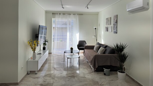 Una sala de estar moderna con un sofá, una mesa de centro y un televisor sobre un mueble bajo. Hay una planta en el suelo y una ventana con cortinas.