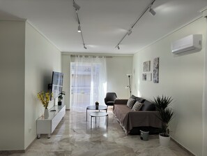 A modern living room with a sofa, a coffee table, and a TV on a stand. There is a plant on the floor and a window with curtains. - Athens Riviera Apartments 300m from Beach (Palaio Faliro)