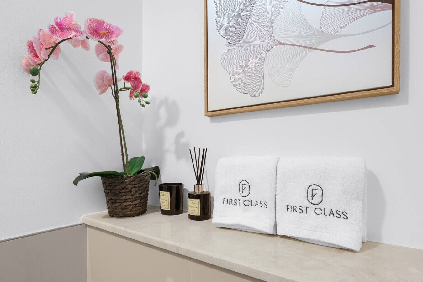 Ein Badezimmer-Waschtisch mit einer Topf-Orchidee, zwei Kerzen und zwei Handtüchern mit der Aufschrift 'First Class'.
