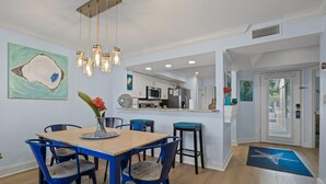 Un comedor con una mesa de madera y sillas azules, una cocina al fondo, y obras de arte en las paredes.