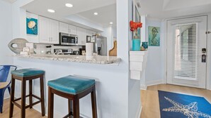 Una cocina moderna con armarios blancos, encimera de mármol y electrodomésticos de acero inoxidable. Hay dos taburetes con cojines color turquesa y una alfombra azul con diseño de estrella de mar.