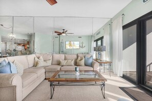 A living room with a sectional sofa, a glass coffee table, a ceiling fan, and a large window with curtains.