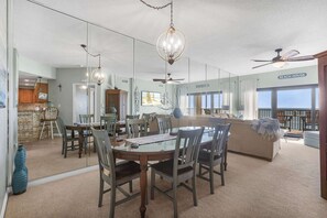 A dining area with a glass table, chairs, and hanging lights. There is a ceiling fan and a sign reading 'BEACH HOUSE'.