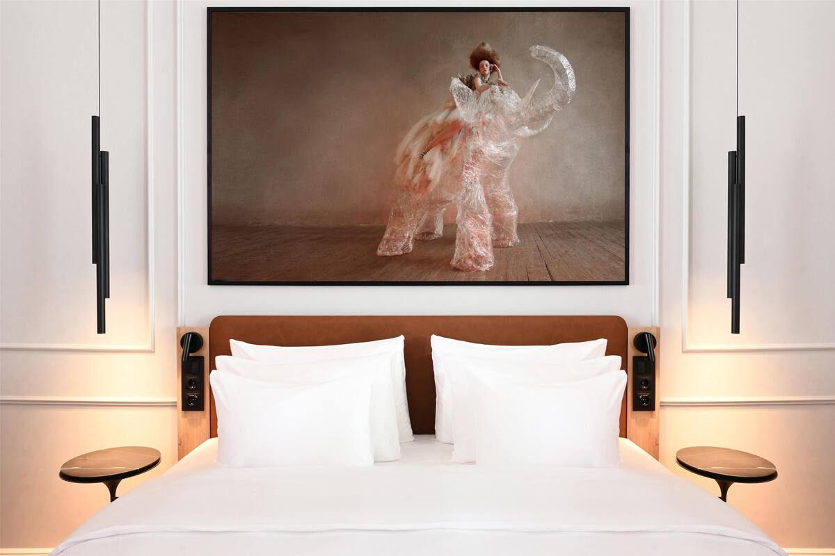 A hotel room with a large bed, two bedside tables, and a painting of a person in a dress on the wall.