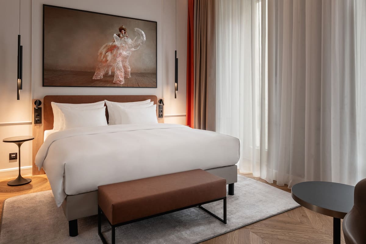 A modern hotel room with a large bed, a bench, a small table, and a painting on the wall.