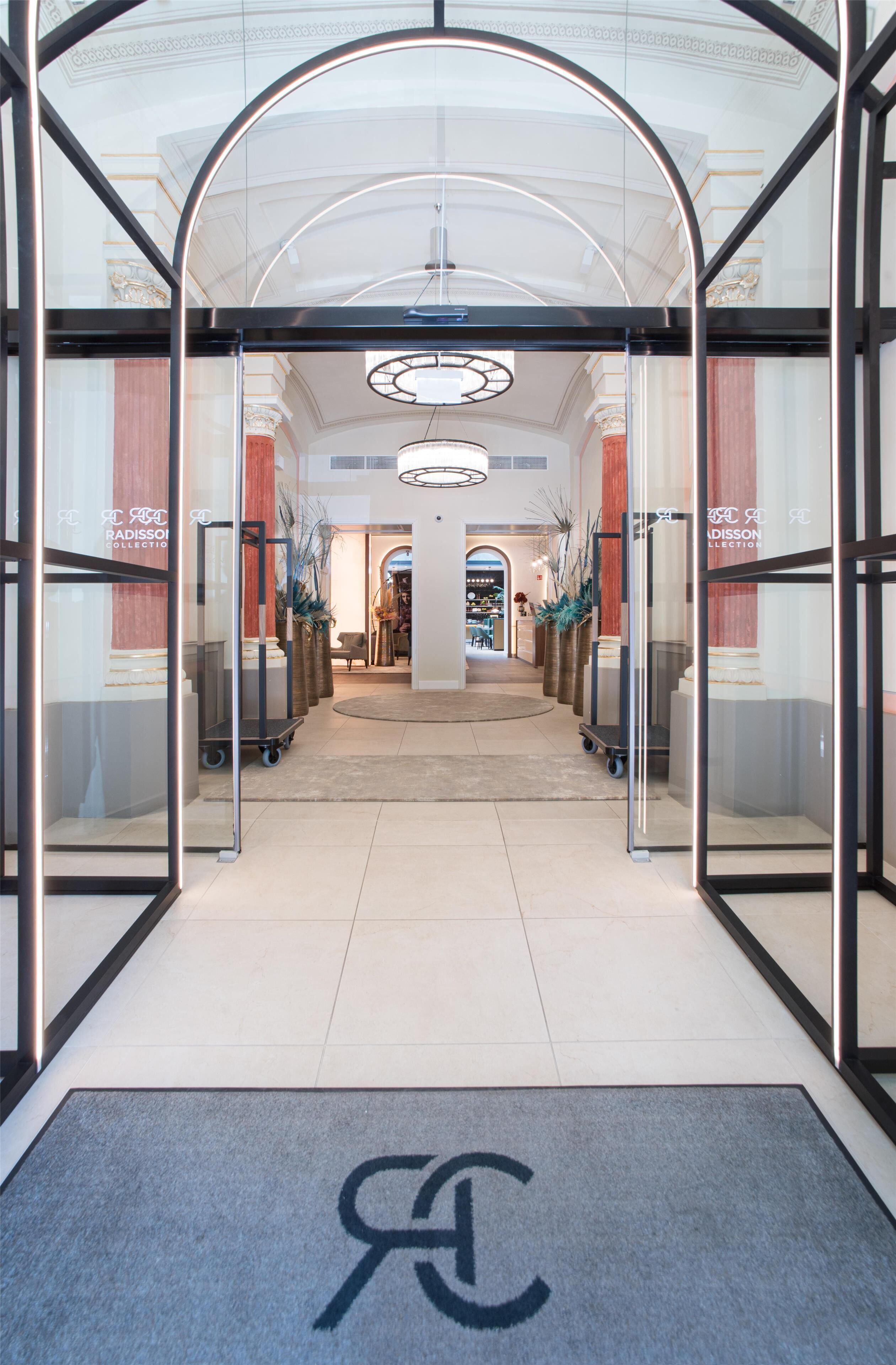 Entrada de un hotel con puerta de cristal, mostrador de recepción y alfombra con un logotipo.