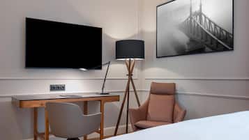 A hotel room with a bed, a desk with a chair, a television, a lamp, and a framed picture on the wall.