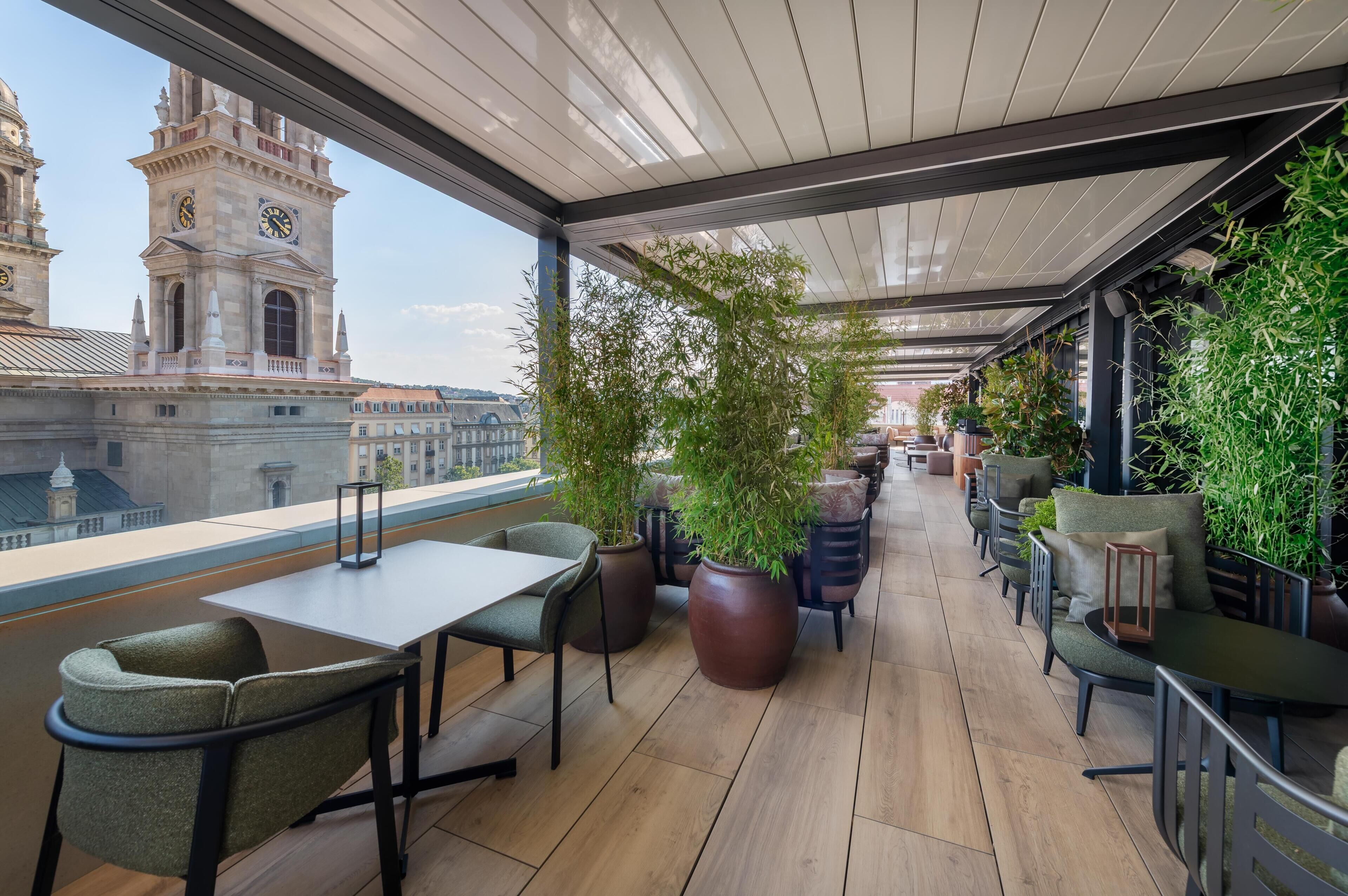 Une terrasse offrant une vue sur des bâtiments historiques, dont un beffroi, aménagée avec des tables et des chaises, et entourée de plantes en pot.