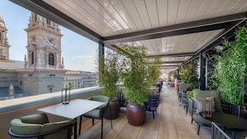 Una terraza con vistas a edificios históricos, incluyendo una torre de reloj, amueblada con mesas y sillas, rodeada de plantas en macetas.