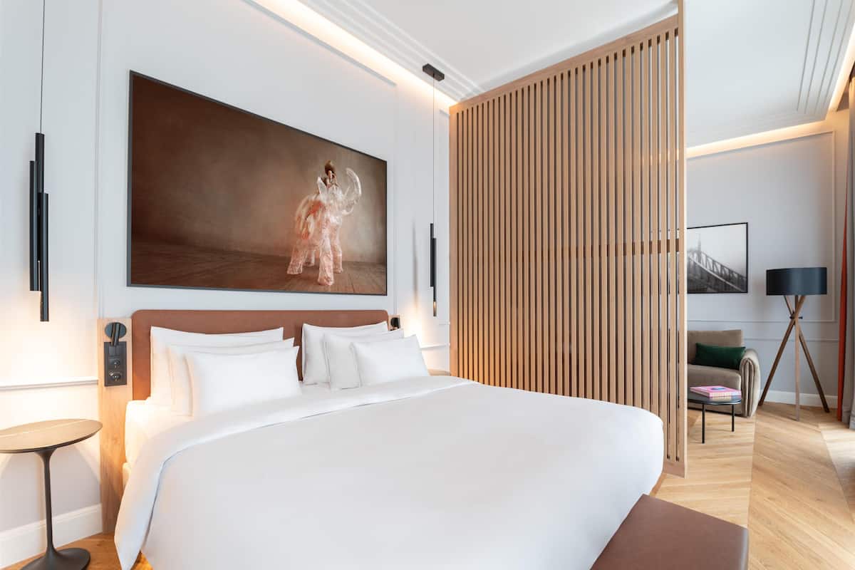 A modern hotel room with a large bed, a wooden headboard, a large painting, a wooden partition, a side table, a floor lamp, a sofa, and a coffee table.