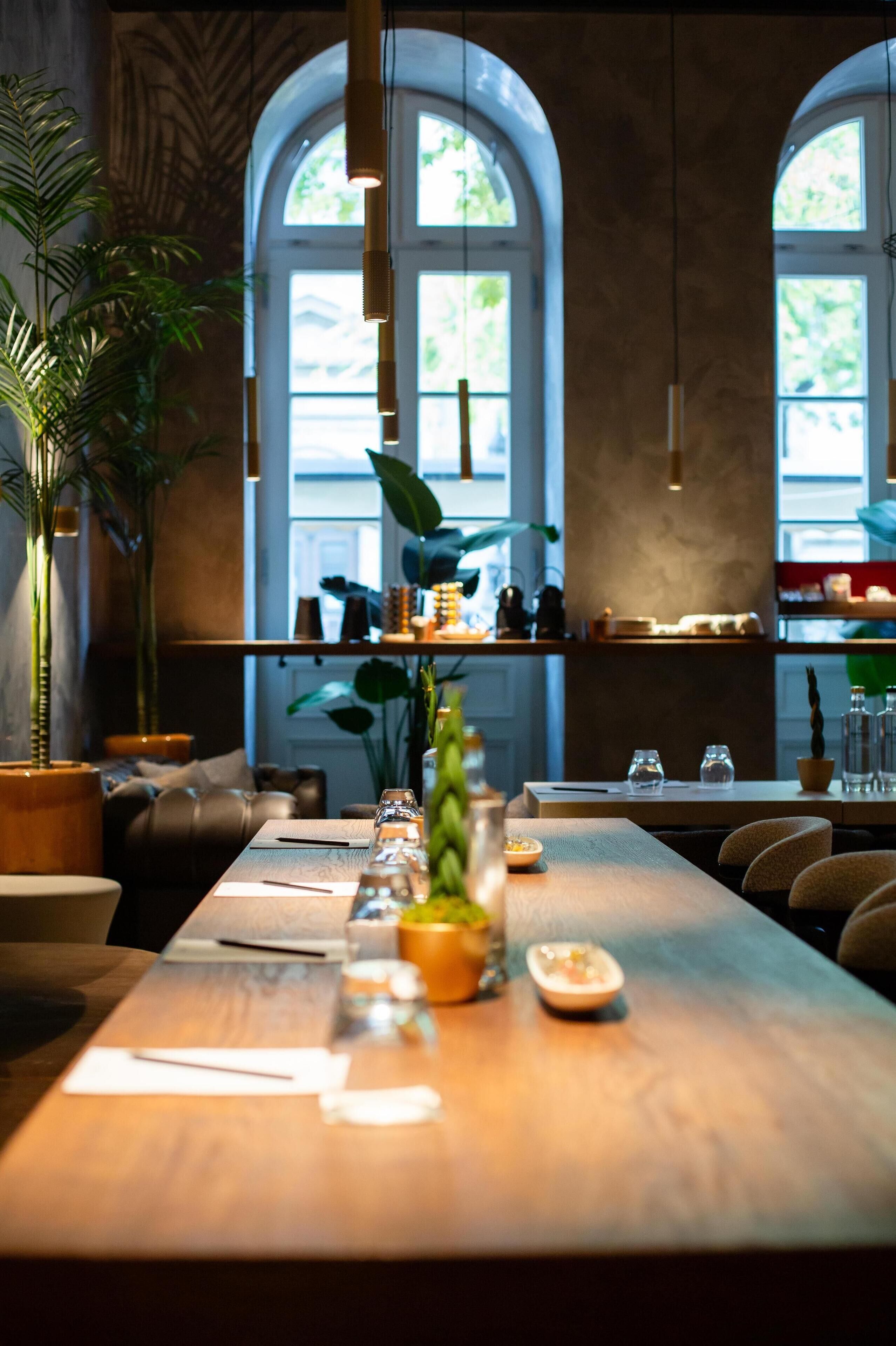 Een restaurant met een lange houten tafel gedekt voor een maaltijd, potplanten en een uitzicht door boogvensters.