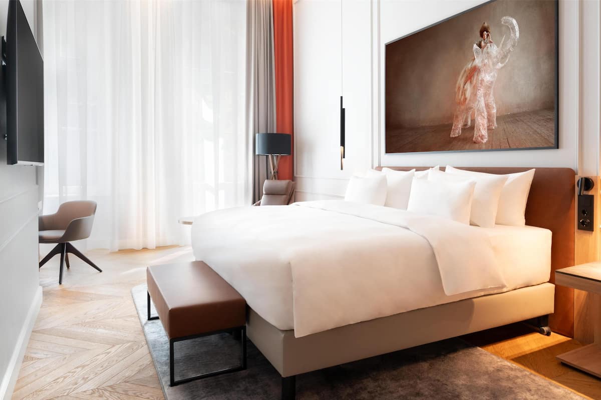 A modern hotel room with a large bed, a desk, a chair, and a large painting on the wall.