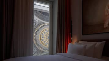 A hotel room with a large bed, a painting on the wall, and a view of a grand architectural structure through a window.