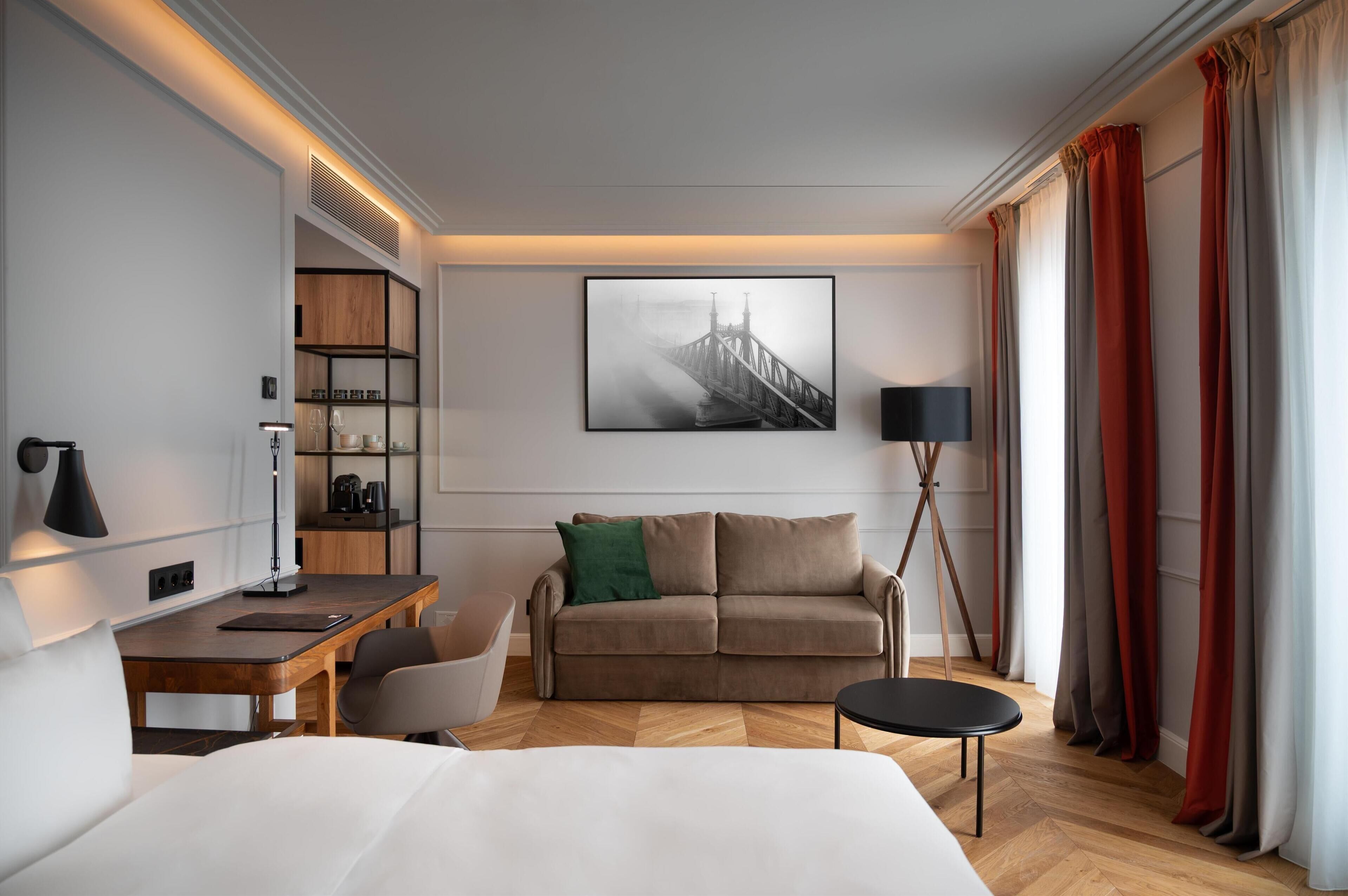 A modern hotel room with a wooden desk, a sofa, a bed, a bookshelf, and a large window with curtains.