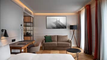 A modern hotel room with a wooden desk, a sofa, a bed, a bookshelf, and a large window with curtains.