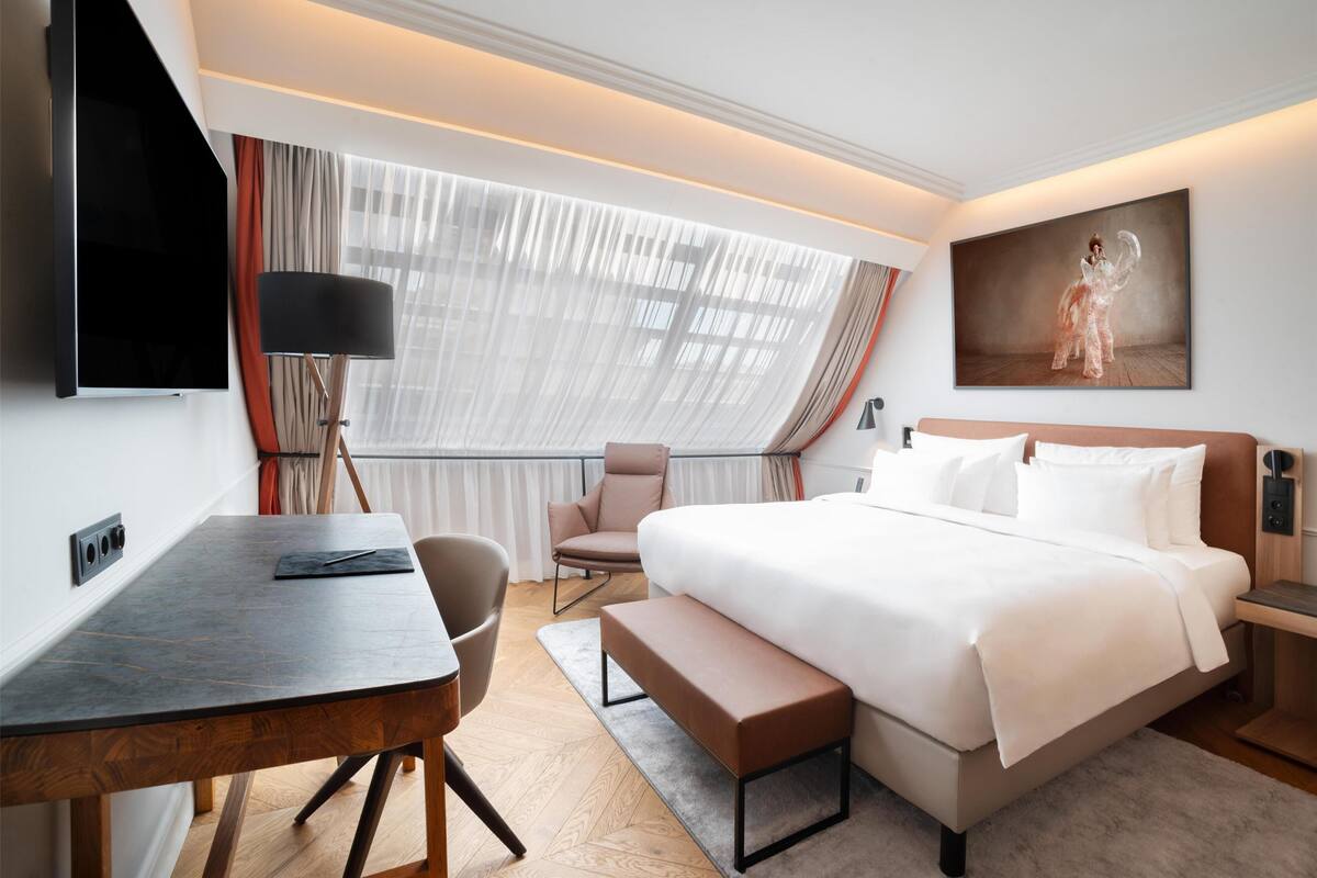 A modern hotel room with a large bed, a desk, a chair, a bench, a TV, and a painting on the wall.