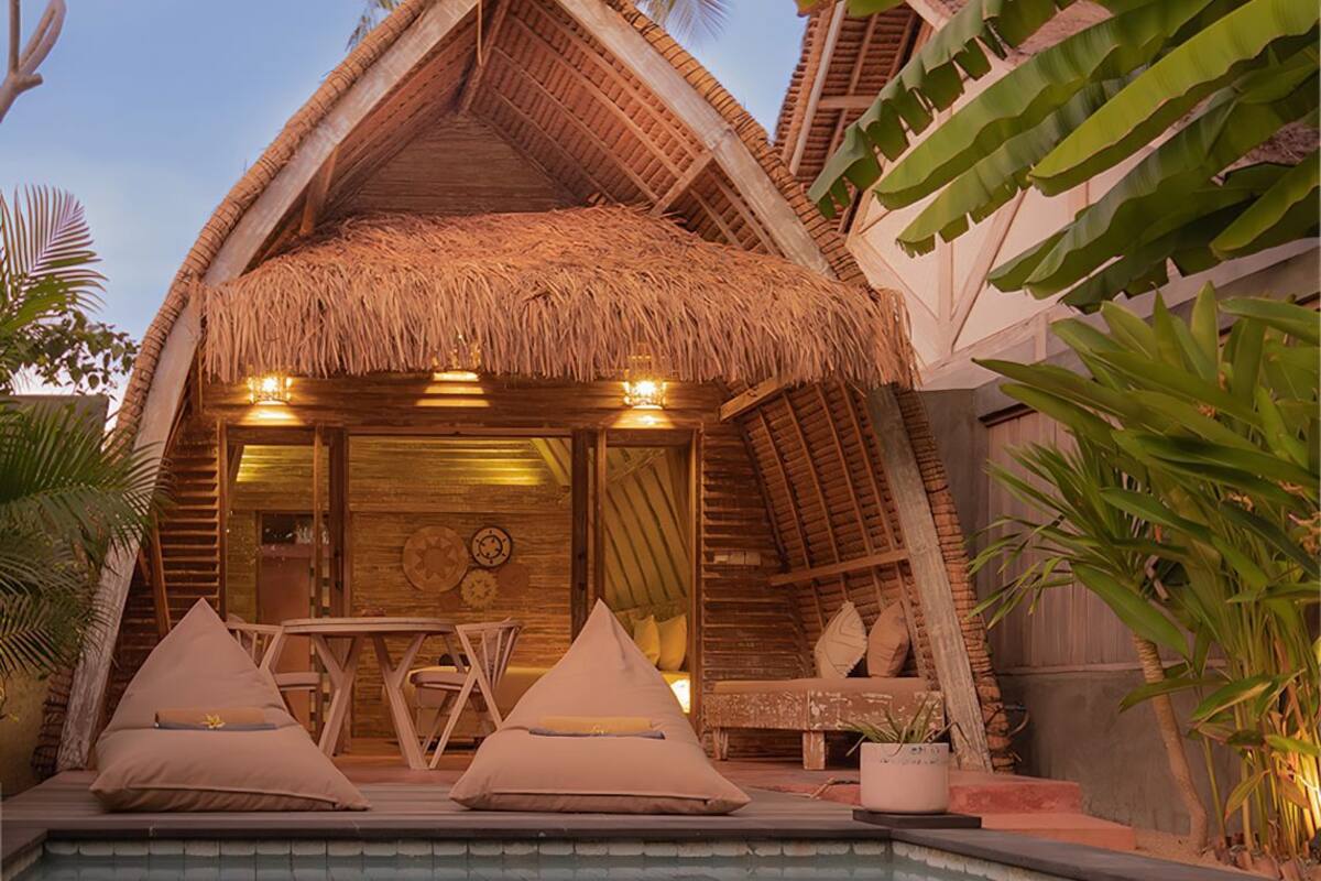 A thatched-roof structure with a pool in front, wooden furniture, and tropical plants.