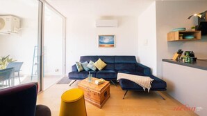 Un salon moderne avec un canapé bleu foncé, une table basse en bois et un pouf jaune. Un tableau orne le mur, et on aperçoit un coin cuisine avec des étagères.