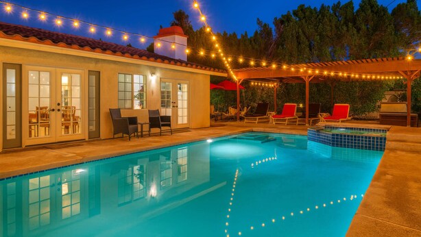 Une piscine illuminée de guirlandes lumineuses, un patio couvert avec des chaises et un bâtiment doté de portes et de fenêtres en verre.