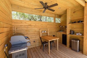 Une cabane en bois équipée d’un ventilateur de plafond, d’un barbecue, d’une table avec des chaises, d’un évier et d’étagères contenant des ustensiles de cuisine.