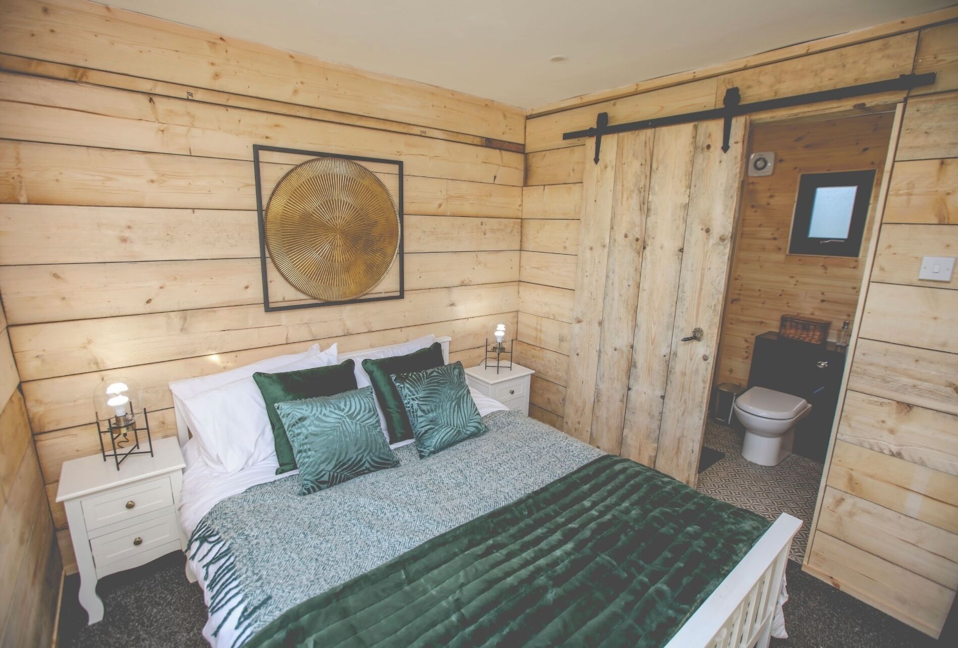 A wooden-paneled bedroom with a bed, two bedside tables, a framed artwork, and a sliding door leading to a bathroom.