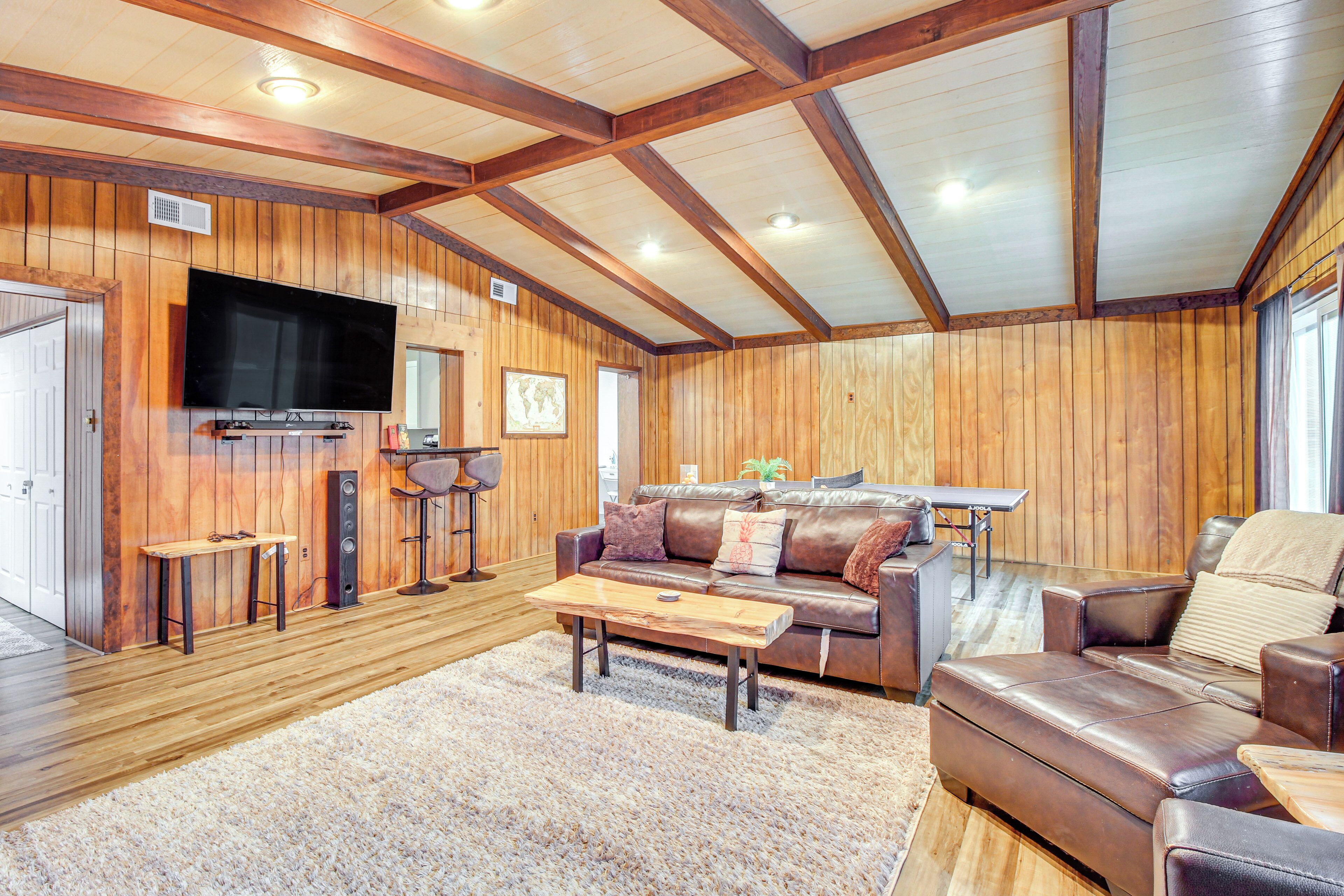 Un vaste salon doté d’un plafond en bois, d’un grand téléviseur à écran plat fixé au mur, d’un canapé en cuir, d’une table basse et d’un coin repas avec une table et des chaises.