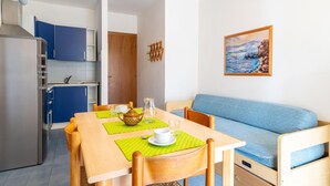 En kompakt opholdsstue med tekøkken, spiseplads med bord og stole, sofa og et maleri af et maritimt landskab på væggen.
