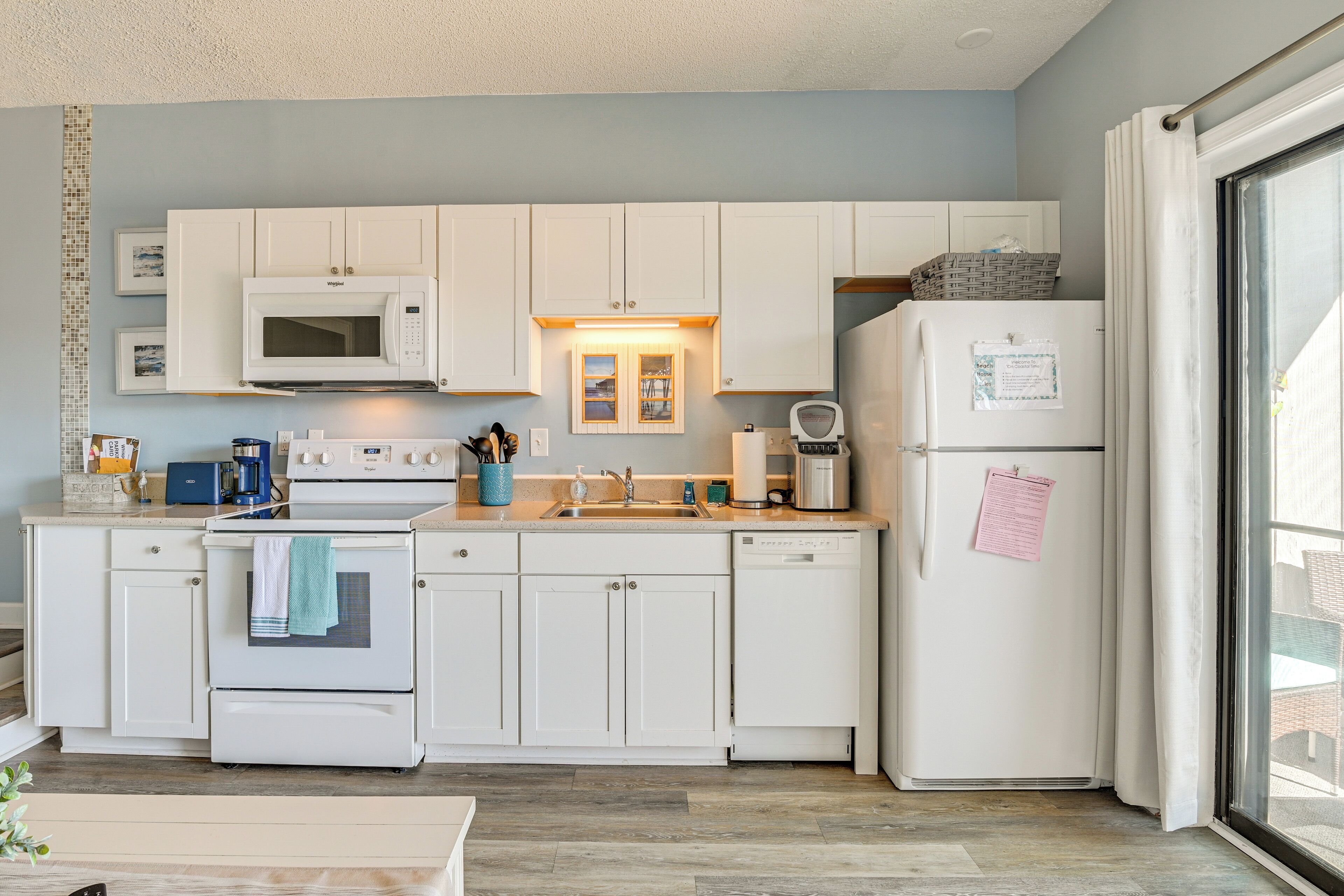 A modern kitchen with white cabinets, a microwave, oven, and dishwasher. There is a window with curtains and a refrigerator with a note attached.
