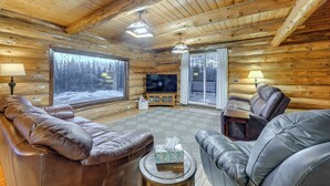 A log cabin interior with a leather sofa, a television, and a large window offering a view of the snowy outdoors.