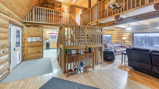 A log cabin interior with a wooden staircase, a living area with a sofa and a coffee table, and a view of the forest through large windows.